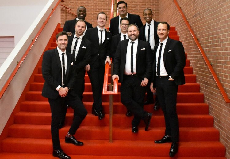 All nine members of Straight No Chaser pose for a photograph on theater stairs after taking photos with fans in Midland, Michigan on Nov. 30, 2024