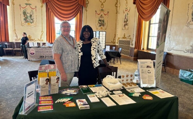 Community health workers Jessica LaGrand, wearing a black dress with a white polka-dot blazer, and Andrew Lowden, wearing a striped button-up shirt with a rainbow lanyard, stand together in front of an informational booth at a health fair.