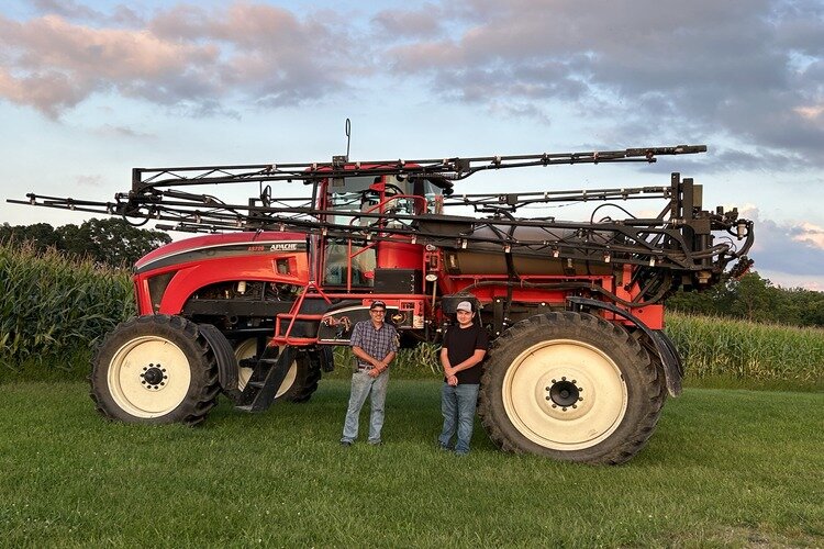 Kevin Dutcher (left), father of Noah Dutcher (right) in front of an Apache self-propelled sprayer.